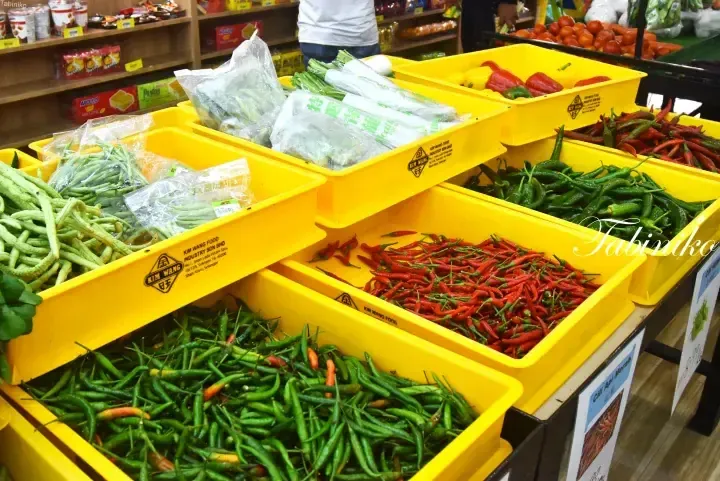Yellow crates filled with fresh green beans, red and green chili peppers, and packaged vegetables at a Malaysian market.