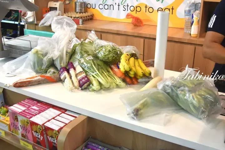 Fresh vegetables and bananas on a checkout counter with Pocky boxes below and a “you can’t stop to eat” sign in the background.