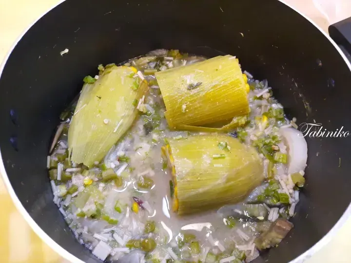 Three corn husk-wrapped tamales simmering in a savory broth with chopped vegetables inside a black cooking pot.