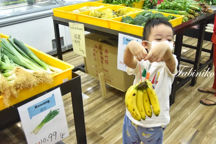 黄色い野菜の箱と価格表示があるマレーシアのスーパーマーケットで、男の子がバナナを持っている様子。