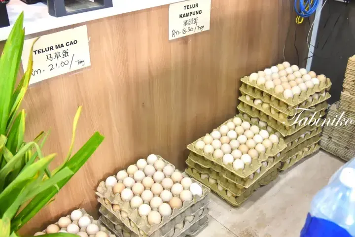 Stacks of egg trays labeled Telur Ma Cao and Telur Kampung at a market stall with price tags in Malaysian Ringgit.