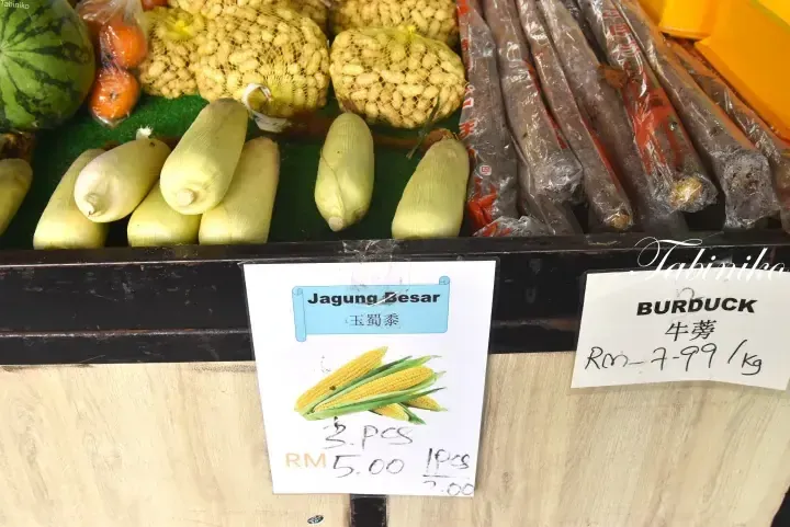 Fresh corn and burdock roots displayed at a market stall with price signs showing Jagung Besar and Burduck prices in Malaysian Ringgit.