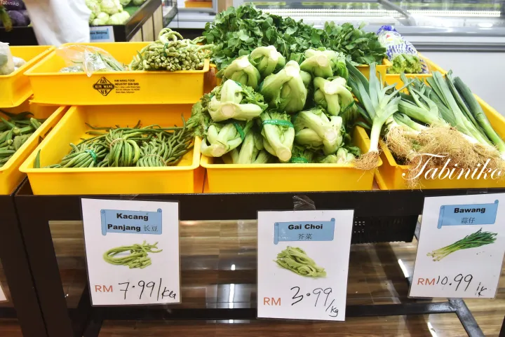 マレーシアの市場で黄色い箱に並べられたKacang Panjang、Gai Choi、Bawangなどの新鮮な野菜と価格表示。