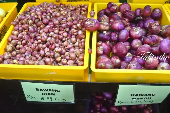 Yellow crates of shallots and red onions at a Malaysian market with price tags showing Bawang Siam and Bawang Merah.