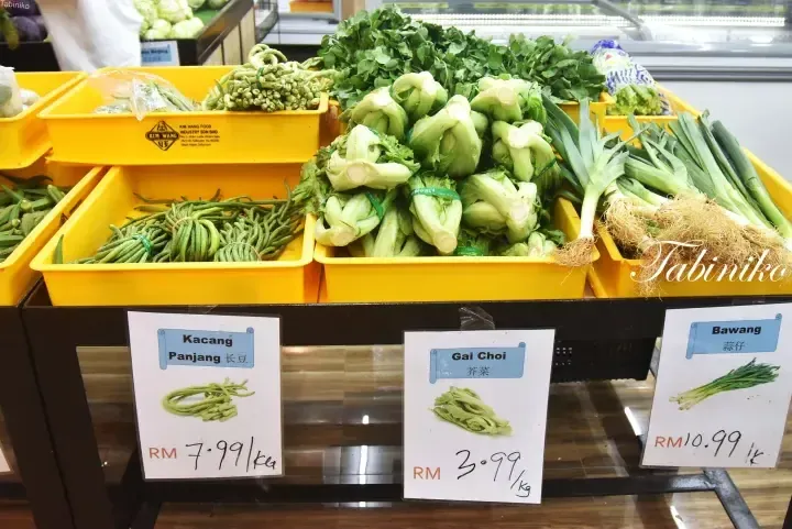Fresh vegetables like Kacang Panjang, Gai Choi, and Bawang displayed in yellow bins with price tags at a Malaysian market.