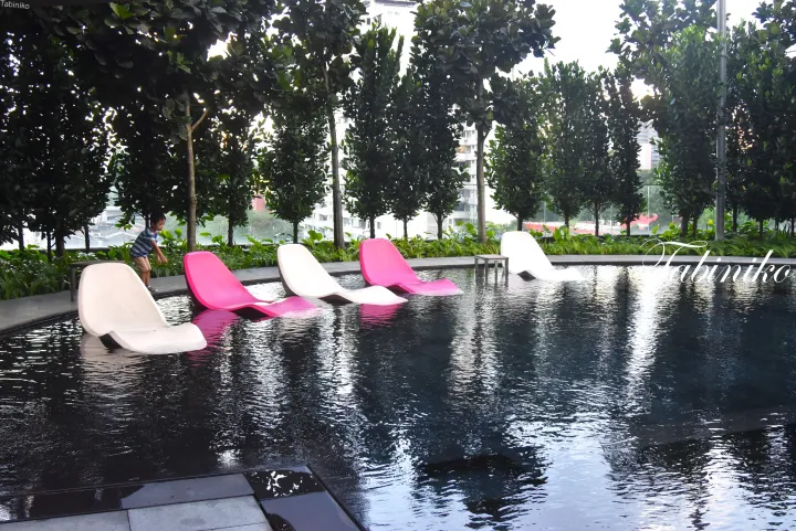 緑豊かな屋上プールに浮かぶピンクと白のラウンジチェア、The Pearl Kuala Lumpur。