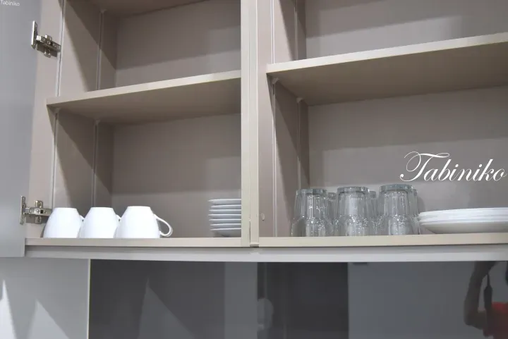 Open kitchen cabinet with shelves holding white mugs, plates, and glasses, featuring the Tabiniko logo.