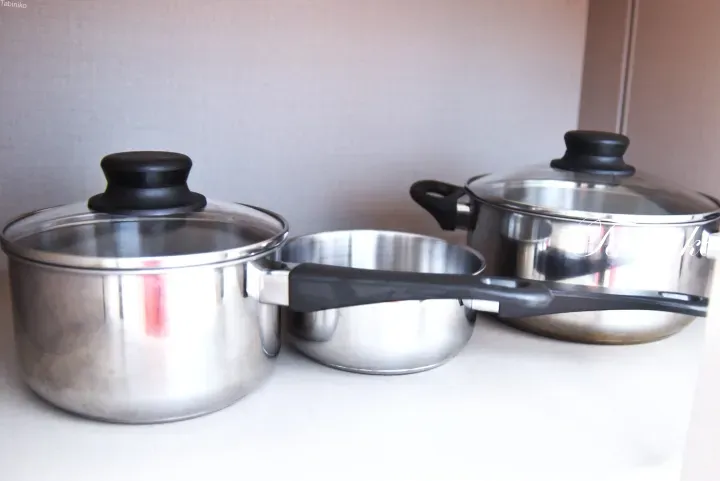 Three stainless steel pots with glass lids and black handles arranged on a white kitchen countertop.