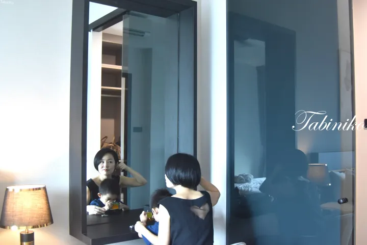 ホテルの部屋で、モダンな化粧台の鏡を見つめる女性と子供。ランプやクローゼットが見える。