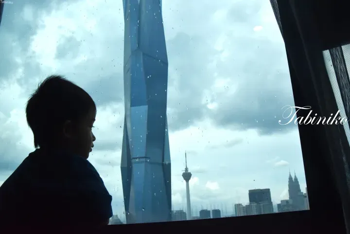 Child looking out a rainy window at Merdeka 118, KL Tower, and Petronas Twin Towers in Kuala Lumpur.