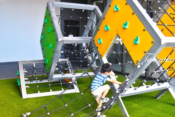 A young boy climbing a modern geometric play structure with rope nets and colorful panels at a property playground.