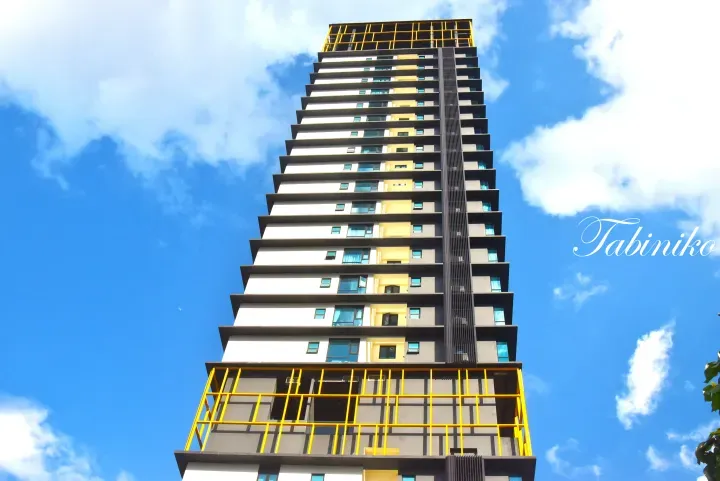Modern high-rise building with yellow accents and balconies against a blue sky, featuring the Tabiniko watermark.