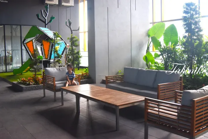 A child sits on a sofa in a modern lounge area with wooden furniture, surrounded by a colorful indoor playground and lush greenery.