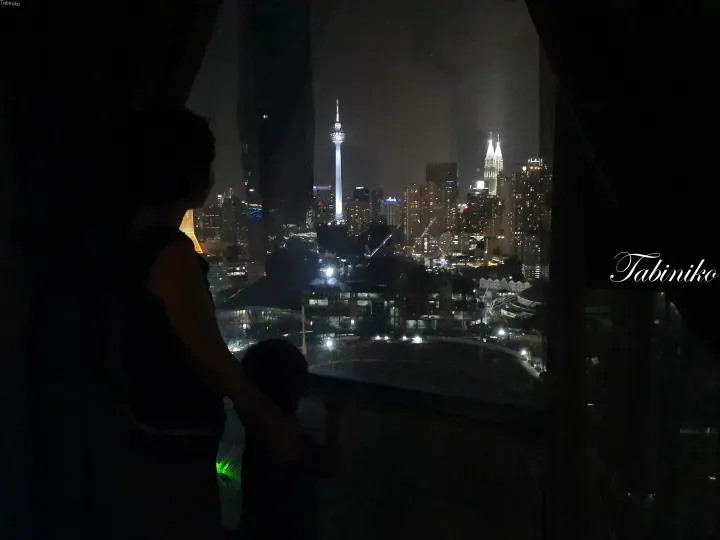 Silhouette of a person and child gazing at the Kuala Lumpur skyline featuring the KL Tower and Petronas Twin Towers at night.
