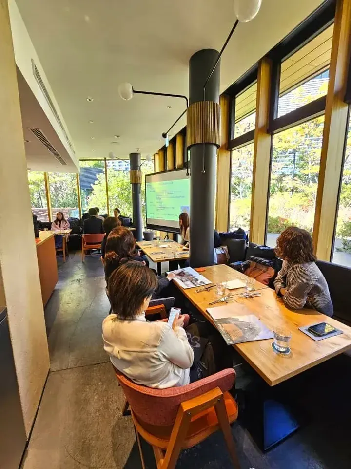 Attendees seated at wooden tables watch a presentation screen in a bright cafe with large windows overlooking greenery.