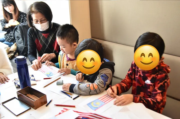 大人が見守る中、子供たちが色鉛筆でマレーシアの国旗を描いている様子。