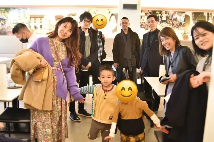 Group of adults and children posing together in a cafe with wall murals, featuring a woman in a purple cardigan and floral dress.