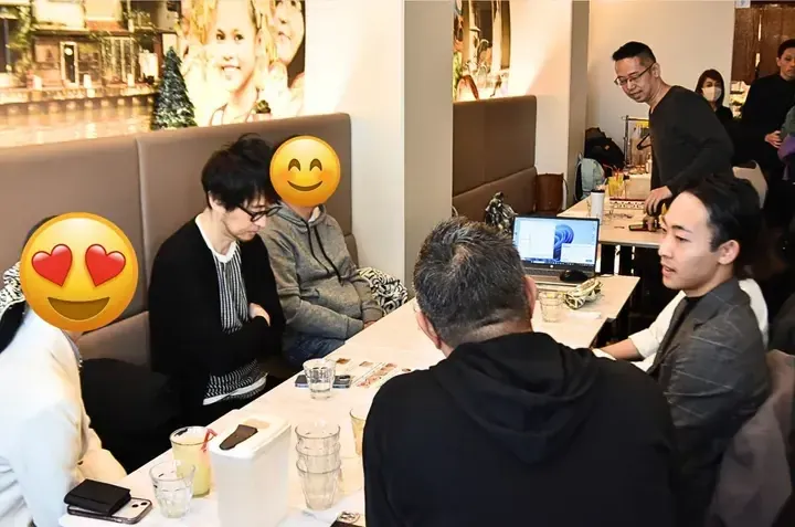 Group of people socializing at a cafe table with a laptop and drinks, featuring wall art of a canal scene.