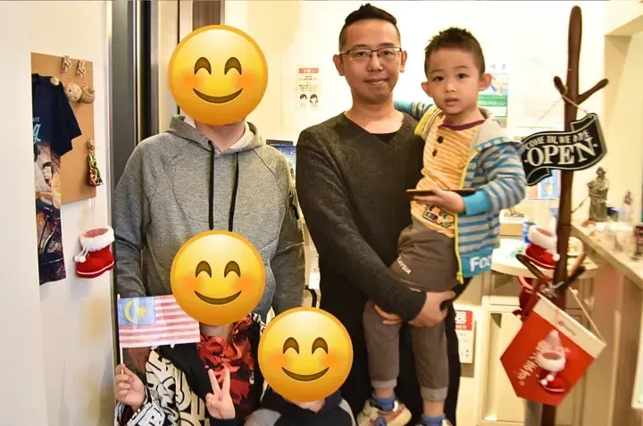 A family poses indoors with a Malaysian flag, standing near a coat rack with an OPEN sign.
