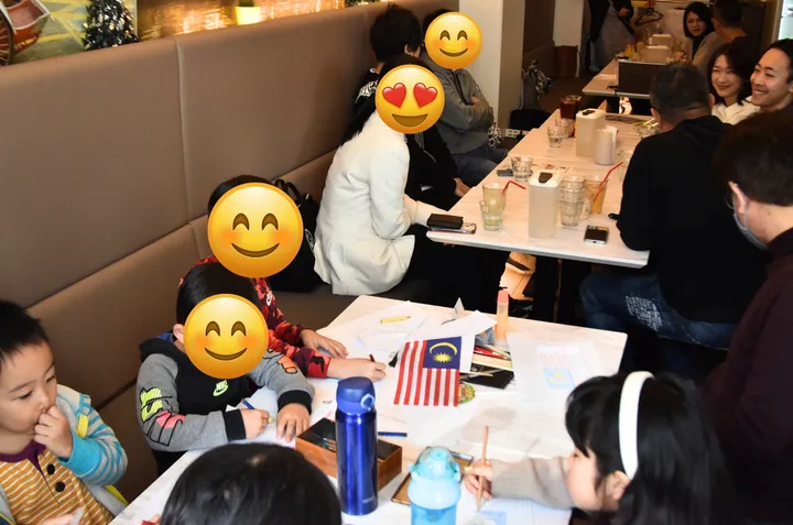 家族の集まりで、マレーシアの国旗を前に子供たちがテーブルで絵を描いている。
