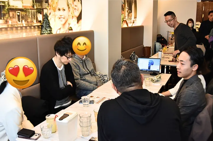 カフェのテーブルでノートパソコンや飲み物を囲み会話する人々のグループと、壁の運河のイラスト。
