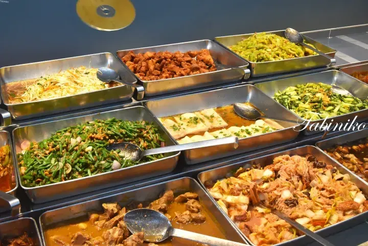 Assorted Malaysian dishes served in stainless steel trays at a buffet, including stir-fried vegetables, tofu, and meat.