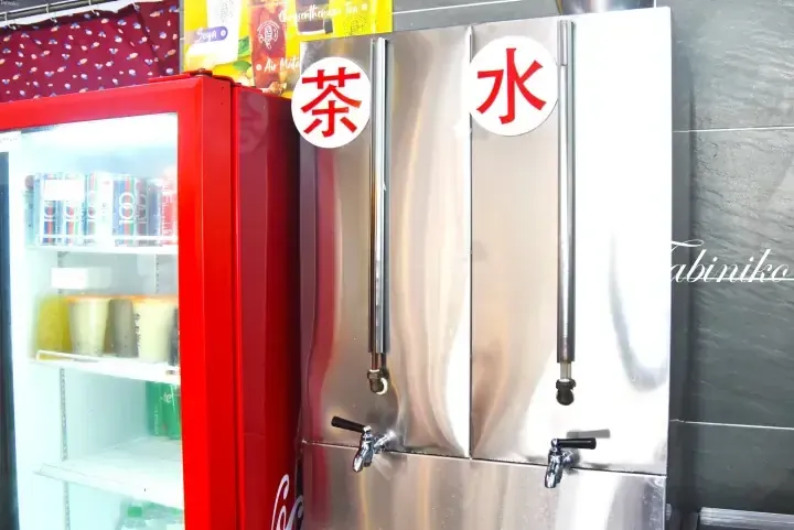 Stainless steel hot and cold water dispenser with Chinese characters for tea and water next to a red beverage refrigerator.