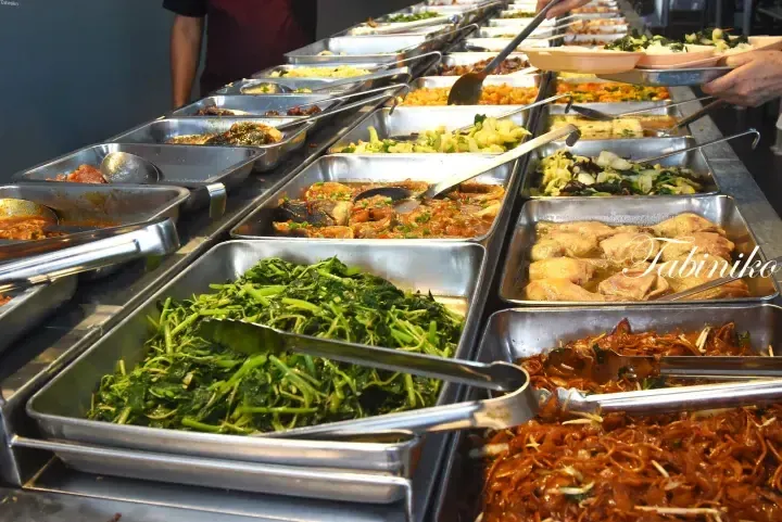 Variety of Malaysian dishes served in stainless steel trays at a buffet, including stir-fried vegetables and noodles.