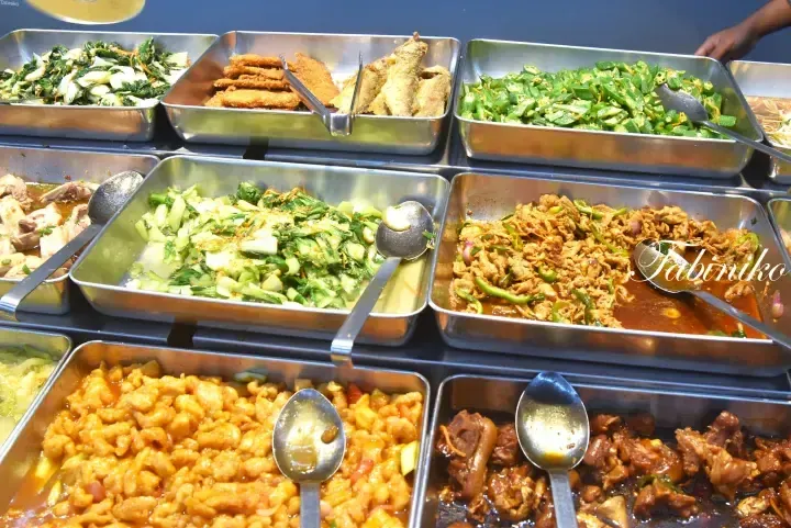 Buffet-style food trays featuring stir-fried vegetables, fried fish, and various meat dishes at a Malaysian eatery.
