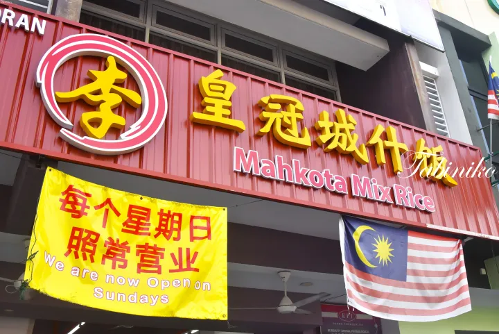 日曜日の営業を告知する黄色いバナーとマレーシア国旗が掲げられた「李皇冠城什饭」の赤い店舗看板。