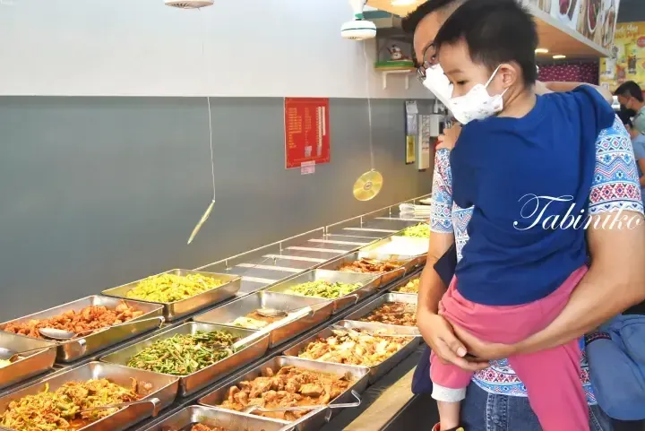 A father holds his child while choosing from a variety of dishes at a Malaysian food stall buffet.
