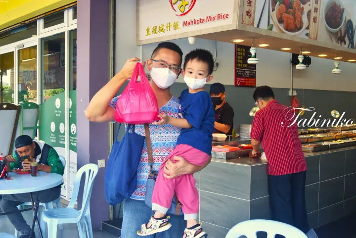 マレーシアの屋台「Mahkota Mix Rice」の前で、テイクアウトの袋を見せる父親と子供。