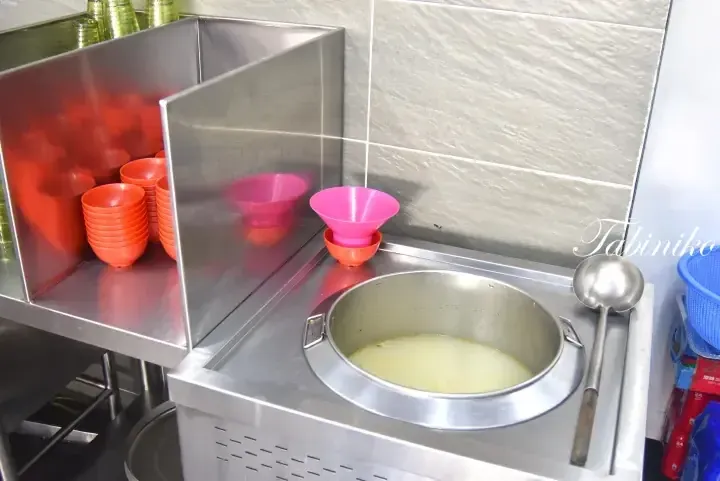 Stainless steel food station with a large pot of soup, ladle, and stacks of colorful bowls in a kitchen.