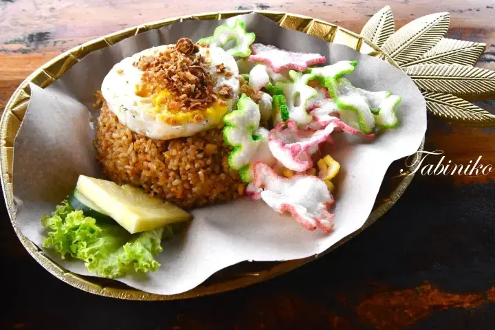 Nasi Goreng Kampung topped with a fried egg and crispy shallots, served with prawn crackers and cucumber on a gold leaf-shaped plate