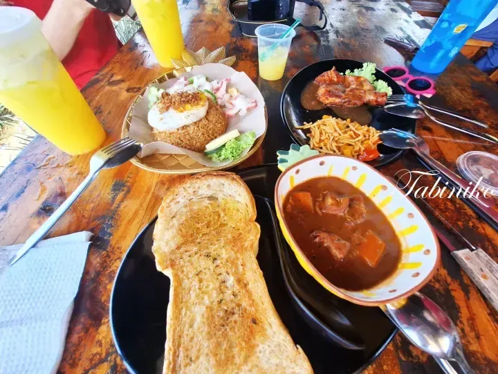 A table spread featuring Nasi Goreng Kampung, chicken curry with spaghetti, beef stew with toast, and yellow drinks at a Malaysian eatery.