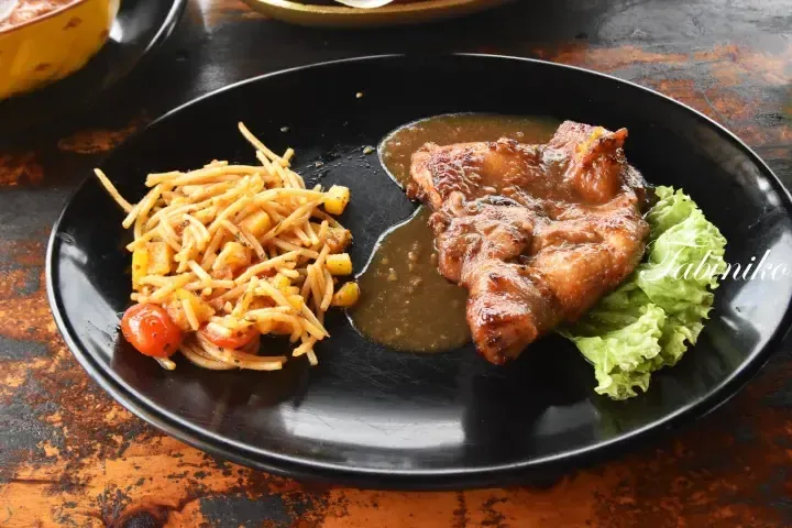 Grilled chicken with gravy and pasta salad served on a black plate at a rustic wooden table.