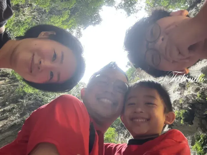 A family of four smiles for a selfie at the entrance of a limestone cave surrounded by lush greenery.