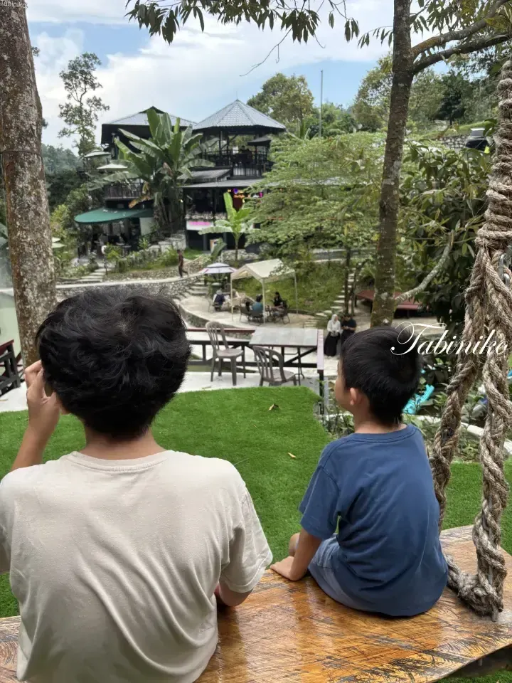 Two boys sit on a wooden swing overlooking a lush hillside resort with traditional architecture and outdoor seating areas.