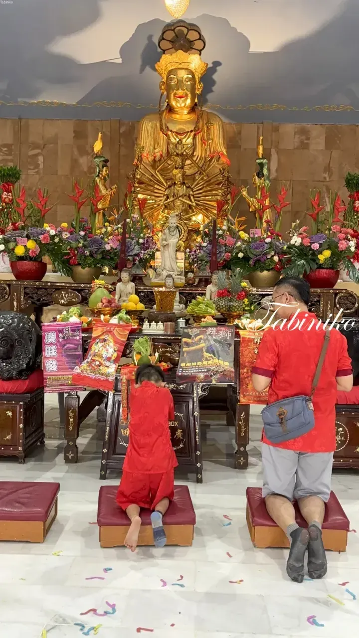 A man and child in red pray before a golden deity statue at a Malaysian temple.