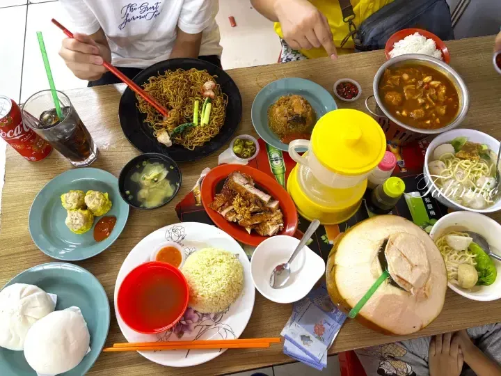 A table spread of Malaysian hawker food including noodles, roasted meat, rice, dim sum, and a fresh coconut.