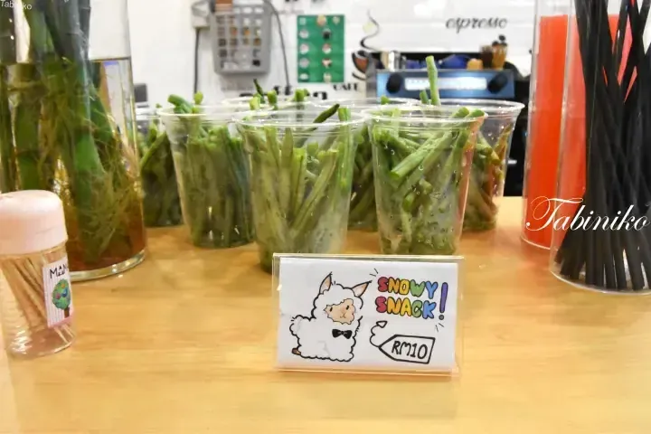Plastic cups of green bean sprouts labeled Snowy Snack RM10 at a cafe counter.