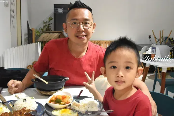 Father and son in matching red shirts enjoy a meal at a restaurant, with the child making a peace sign beside plates of rice and curry.