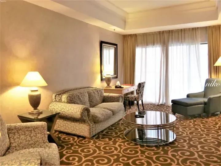 Spacious hotel suite living area with floral sofa, round glass coffee table, and large window with sheer curtains
