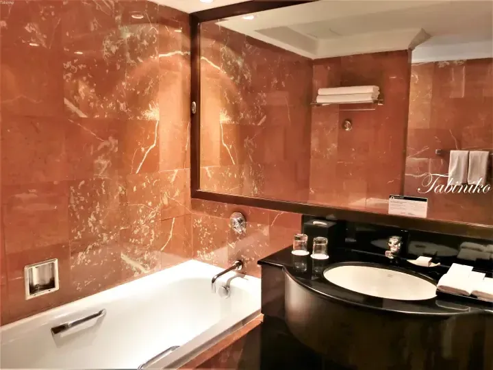 Luxury hotel bathroom with reddish marble walls, a white bathtub, and a black vanity sink.
