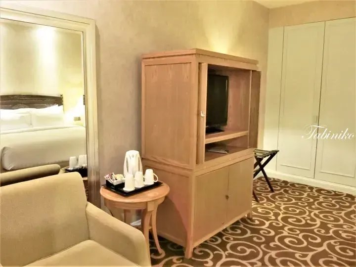 Wooden TV cabinet with sliding doors, tea station, and beige sofa in a hotel room with patterned carpet.