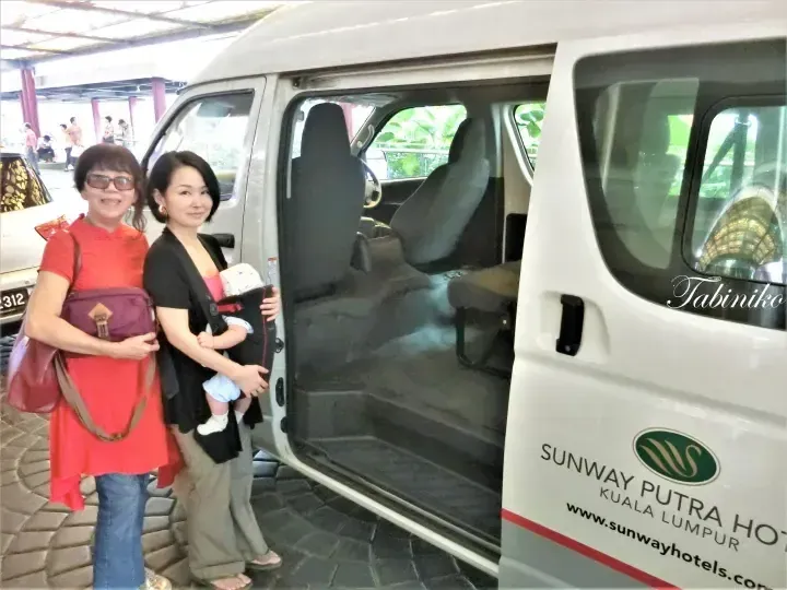 Two women and a baby stand beside a white Sunway Putra Hotel shuttle van with its side door open.