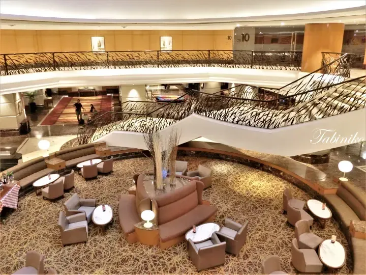 Spacious hotel lobby with curved sofas, marble tables, and a grand staircase with decorative railings.