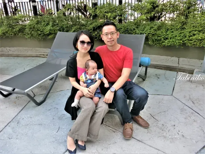 A family of three sitting on a lounge chair at a property rooftop terrace with greenery and city views.