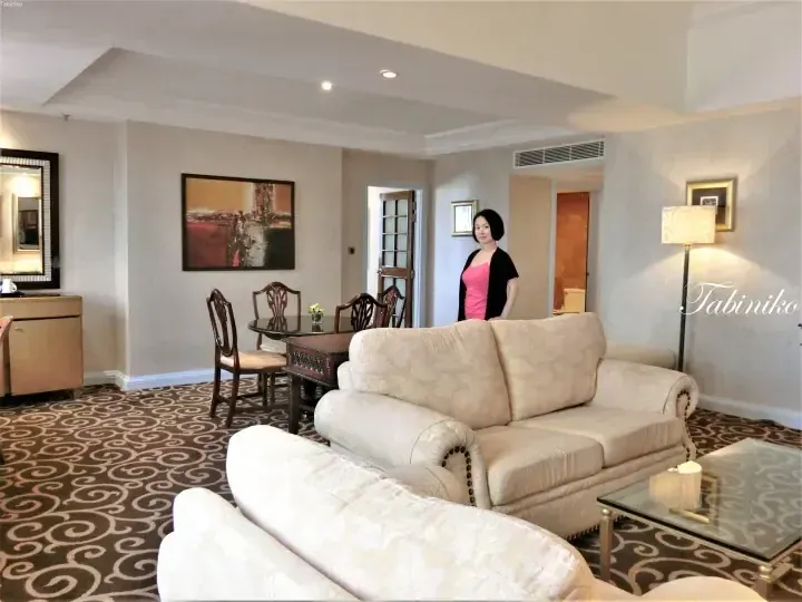 Woman standing in a spacious hotel suite living room with beige sofas, a dining table, and patterned carpet.