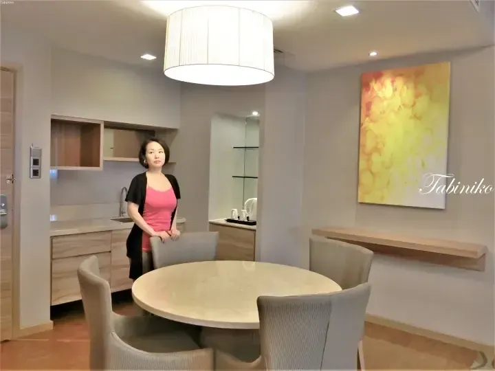 Woman standing in a modern dining area with a round table, kitchenette, and abstract art in a serviced residence.